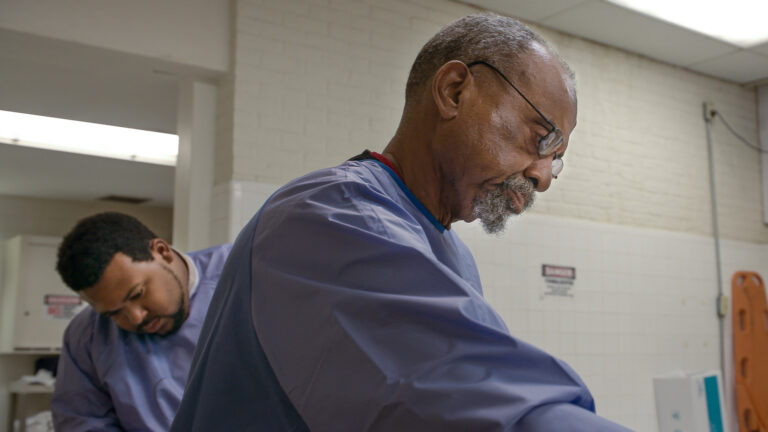 Two medical examiners in scrubs working in a morgue, one with gray hair and glasses.