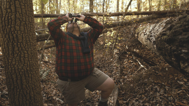 Man birdwatching with binoculars in a dense forest.