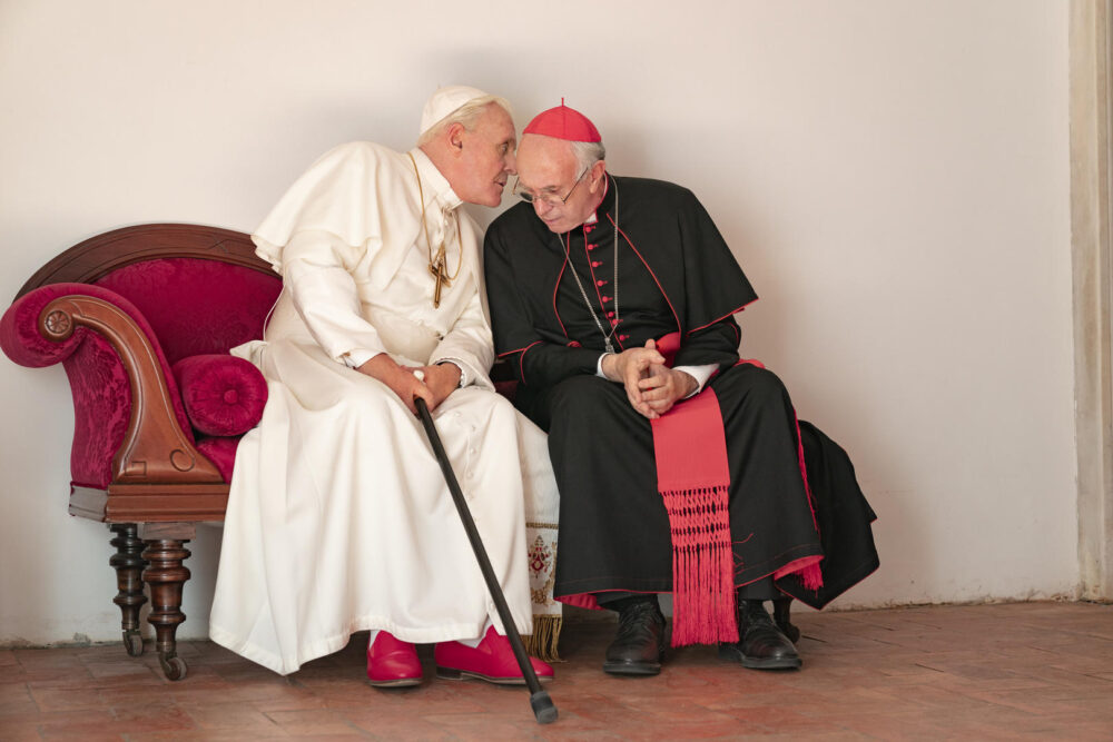 Anthony Hopkins and Jonathan Pryce in "The Two Popes" movie, sitting on a red couch.