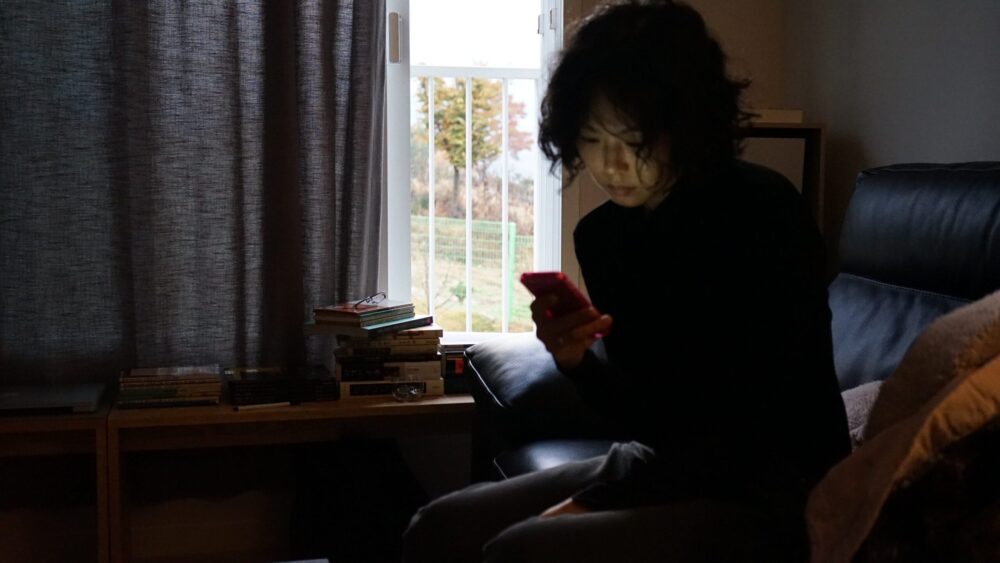 Woman looking at her phone in a dimly lit room by a window.