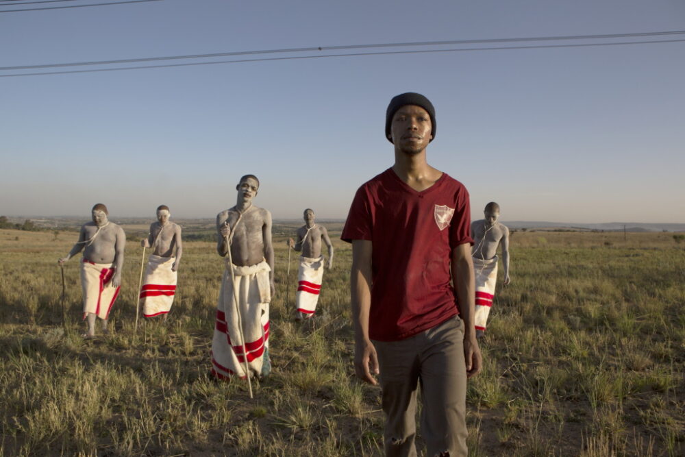 Man in modern clothes stands before initiates in traditional Xhosa attire.