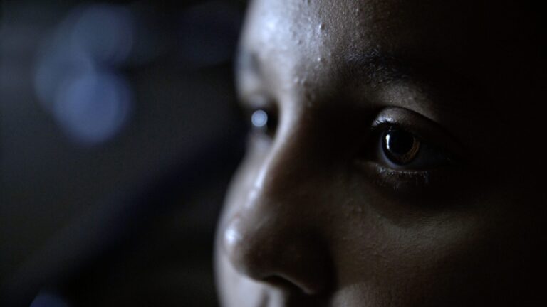 Close-up of a child's face, eyes reflecting light, possibly gazing at the sky.