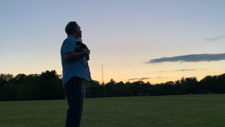 Man with camera silhouetted against sunset sky on empty green field. "There Was Nobody Here We Knew.