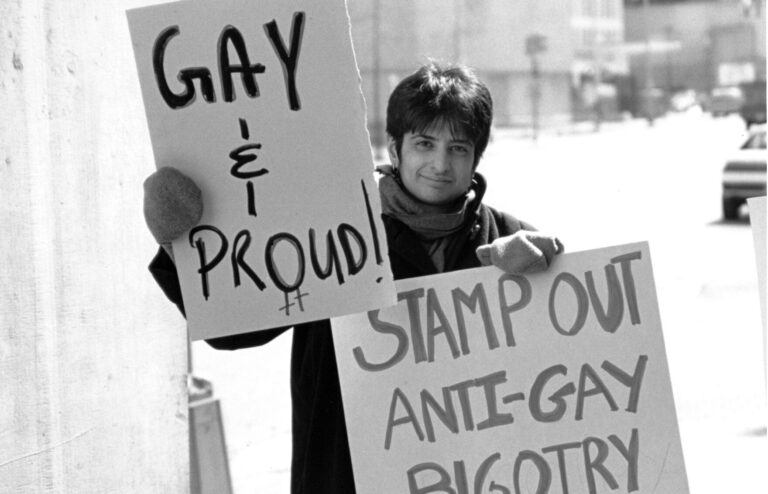 Activist holding signs: "Gay & Proud" and "Stamp Out Anti-Gay Bigotry". LGBTQ rights protest.