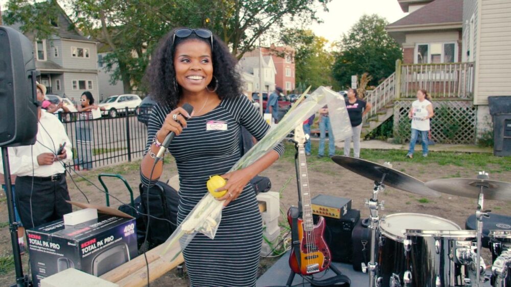Woman with microphone and lulav at outdoor event. "They Ain't Ready for Me" event in background.