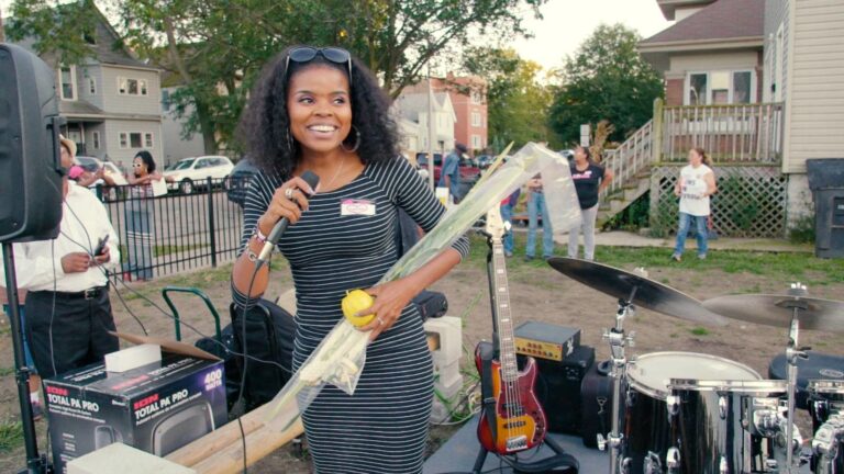 Woman with microphone and lulav at outdoor event. "They Ain't Ready for Me" event in background.