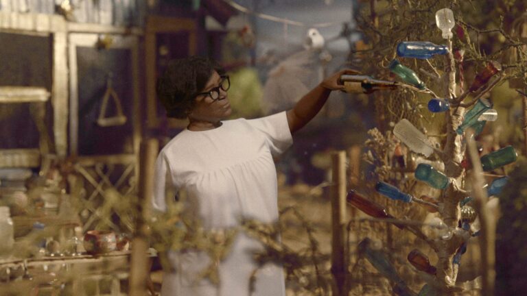 Woman decorates bottle tree in artistic outdoor setting.