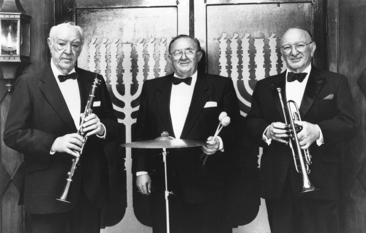 Three musicians with clarinet, cymbals, and trumpet in front of a menorah.
