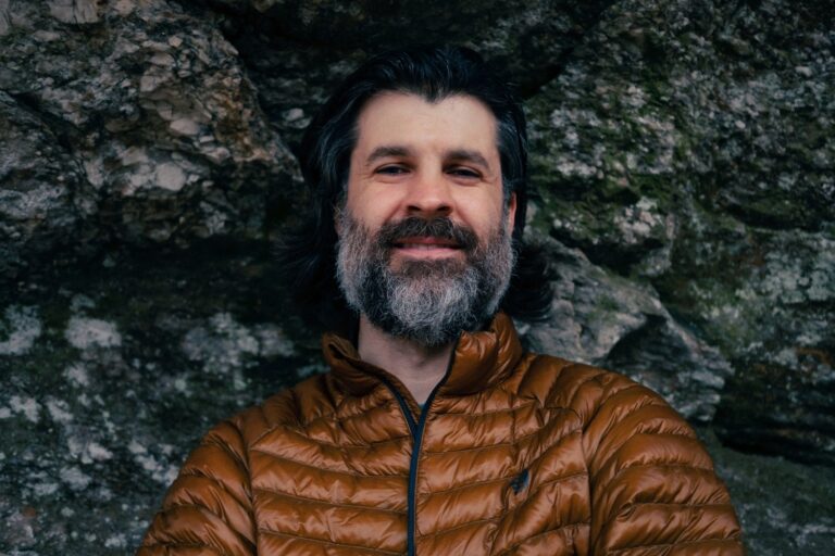Tim Grant wearing a brown puffer jacket, smiling against a rock background.