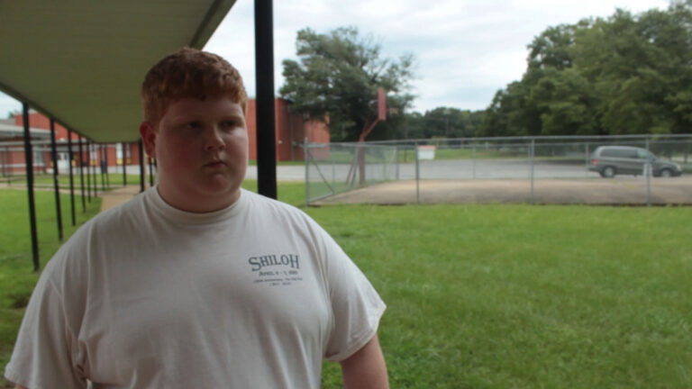 Teen boy in Shiloh shirt stands outside on a school campus.