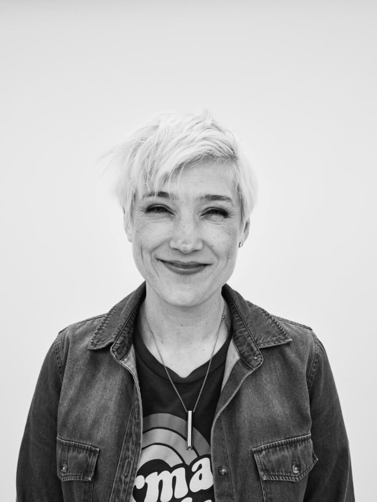 Tracie Laymon, smiling with short blonde hair, wearing a denim jacket and necklace.