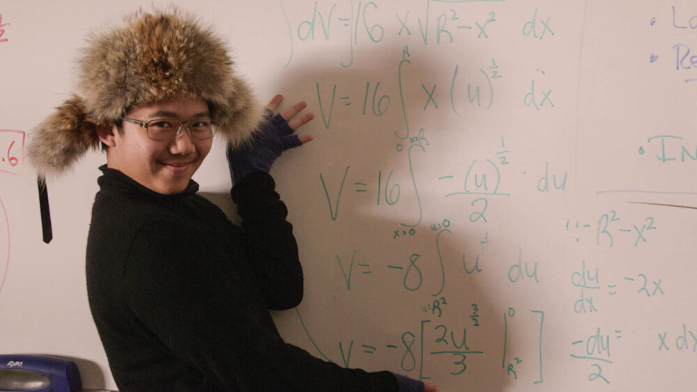 Man in fur hat explaining math on a whiteboard with integral equations.