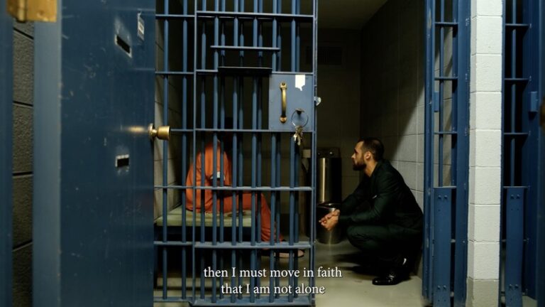 Prison scene: man in orange jumpsuit behind bars, another man kneeling.