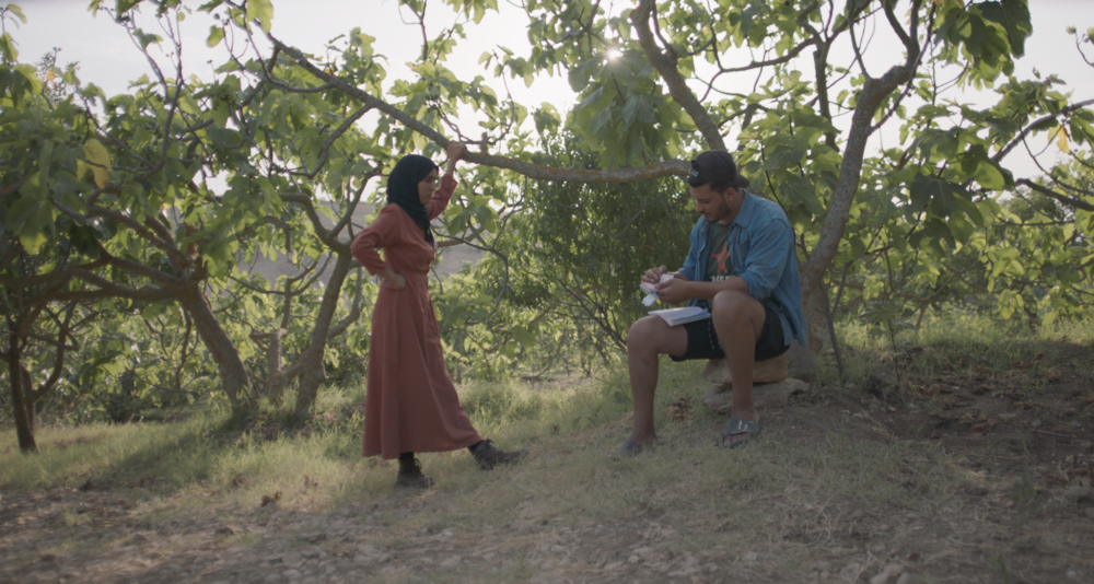 Woman in hijab stands by a man sketching under fig trees.