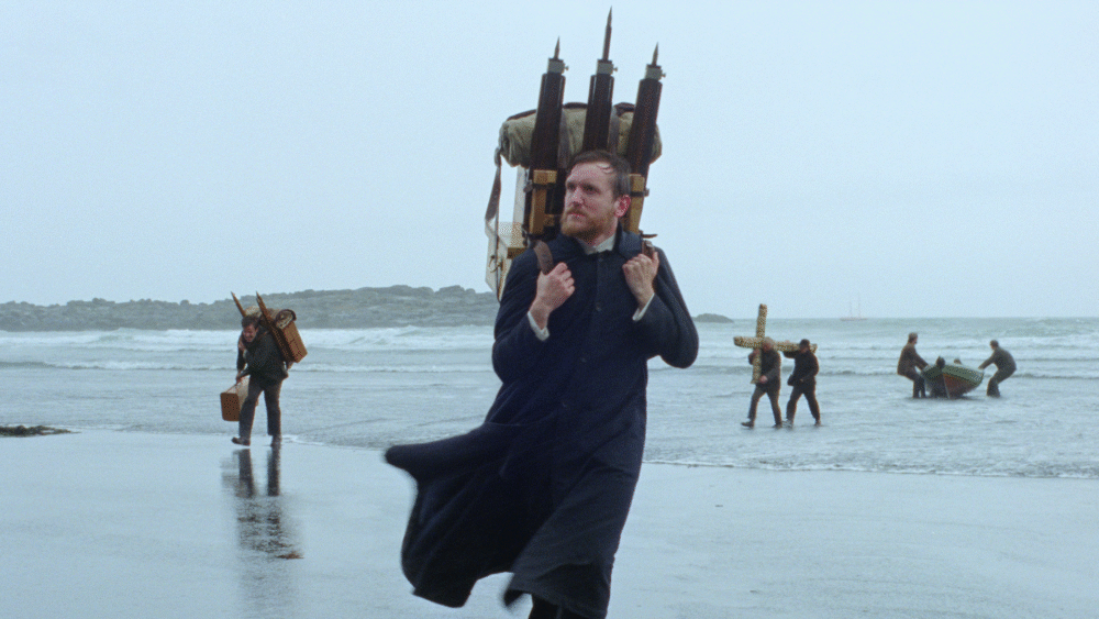 Godland film scene: Priest carrying photographic equipment on a beach.