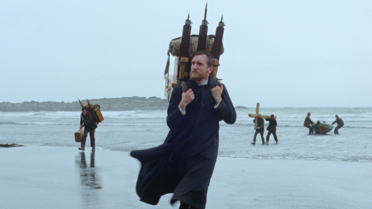 Godland film scene: Priest carrying photographic equipment on a beach.