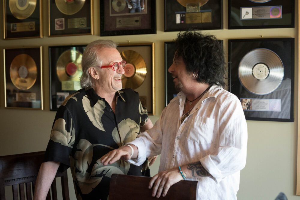 Two musicians laughing in front of gold and platinum record awards.