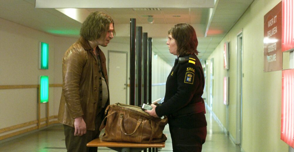 Man questioned by border security, bag on table.