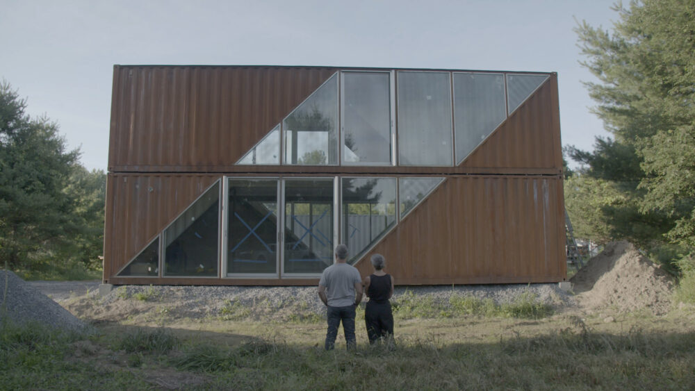 Couple admires a modern shipping container home with triangular windows.
