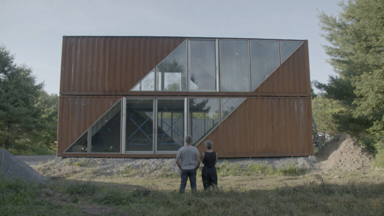 Couple admires a modern shipping container home with triangular windows.