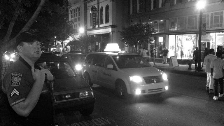 Wilmington police officer stands by car as a Yellow Cab drives by on a city street at night.