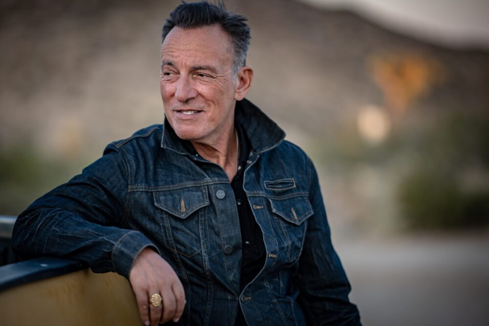 Bruce Springsteen in a denim jacket, gazing into the distance in a desert landscape.