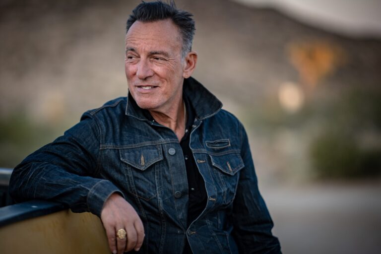Bruce Springsteen in a denim jacket, gazing into the distance in a desert landscape.