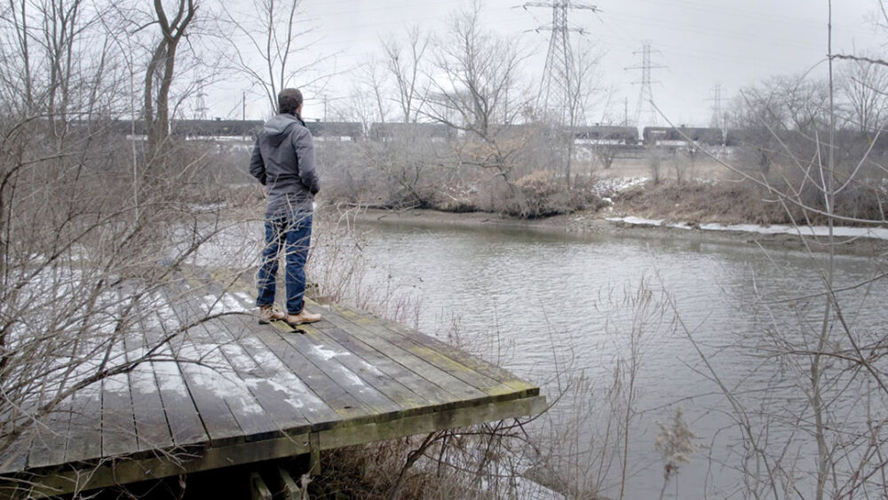 Man looks at a river from a wooden platform, train in background.