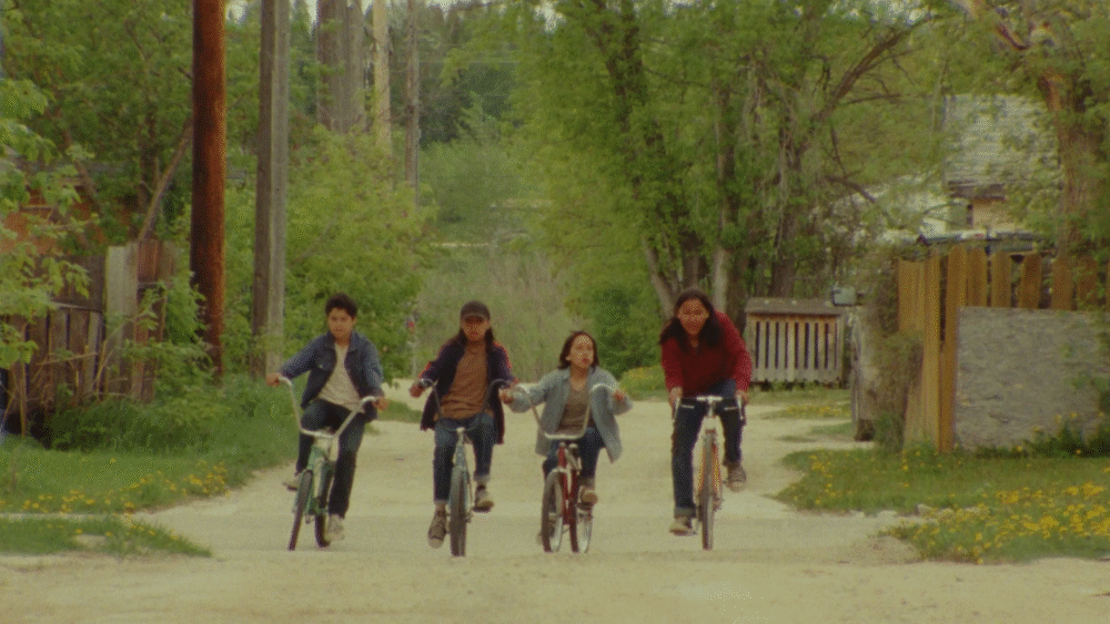 Four Indigenous children ride bikes down a dirt road in a rural setting.