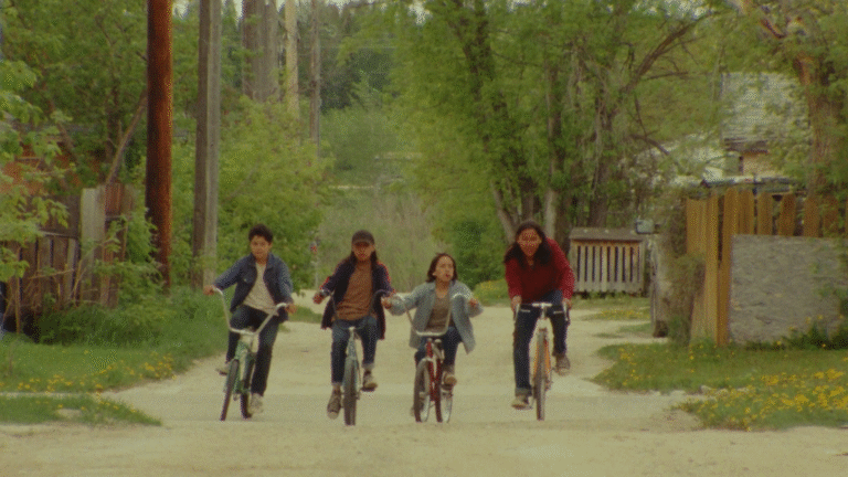Four Indigenous children ride bikes down a dirt road in a rural setting.