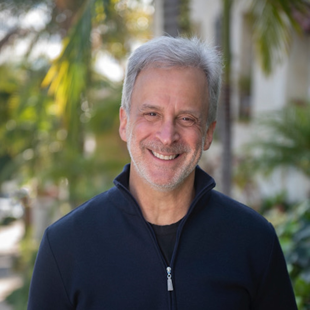 William Goldenberg, smiling in a navy zip-up sweater in front of palm trees.
