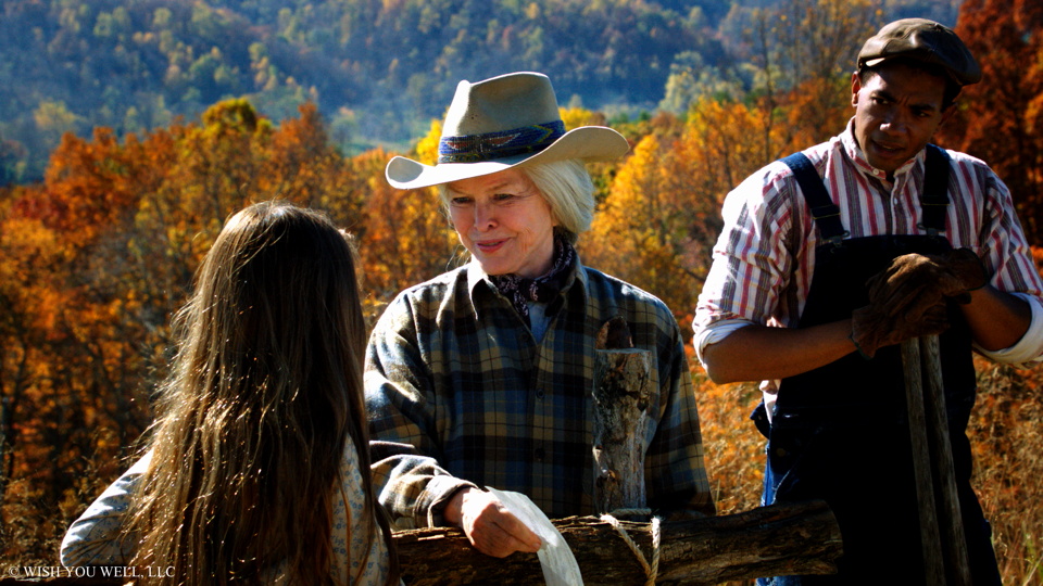 Family scene from "Wish You Well" movie: girl, woman in cowboy hat, man in overalls near a fence with autumn trees behind.