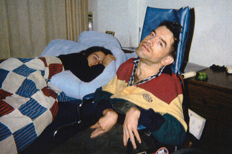Man in wheelchair next to a sleeping woman under a quilt.