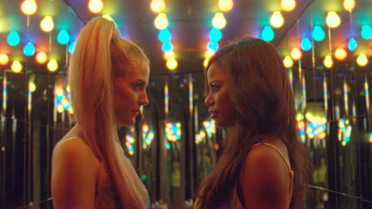 Two women face each other amidst colorful lights and mirrors, reminiscent of the film Zola.