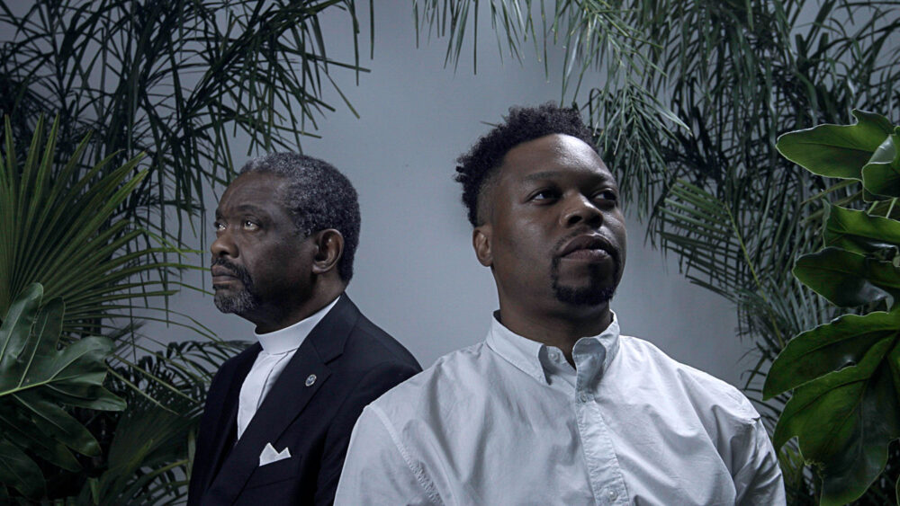 Two men amidst greenery, one in clerical attire.
