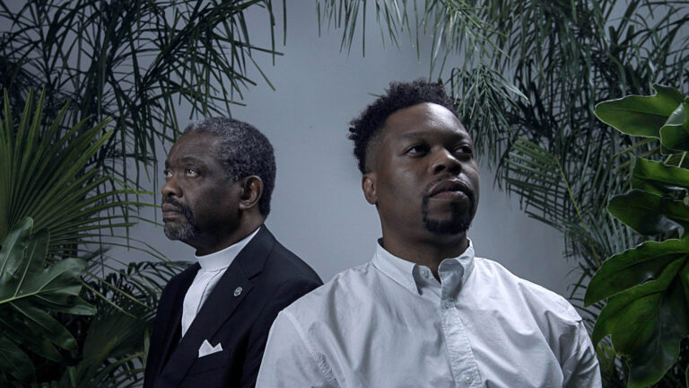 Two men amidst greenery, one in clerical attire.