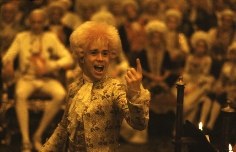 Young Mozart, portrayed in Amadeus, with a powdered wig, excitedly gestures in a crowded room.