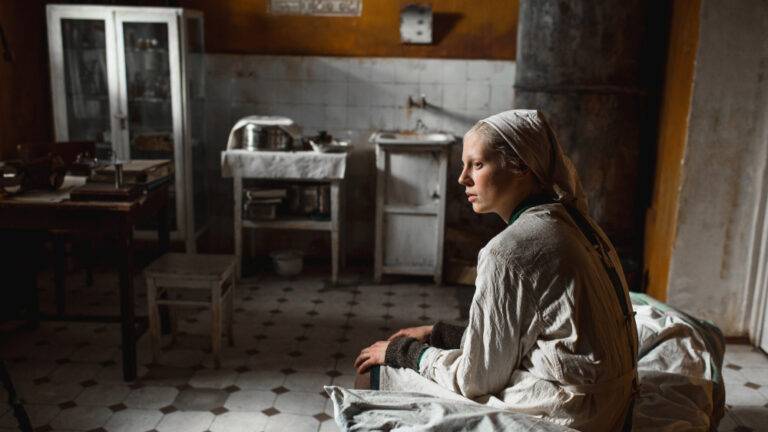 Beanpole film still: A woman sits in a sparse, tiled room looking thoughtful.