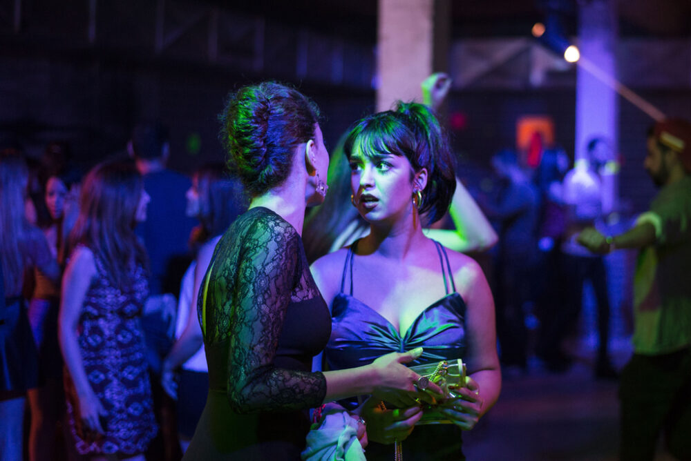 Two women talking at a nightclub, bathed in blue and purple light.