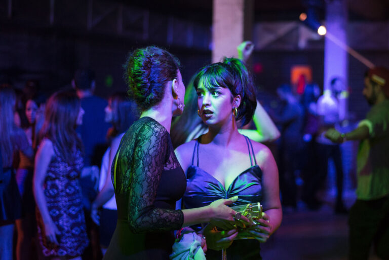 Two women talking at a nightclub, bathed in blue and purple light.