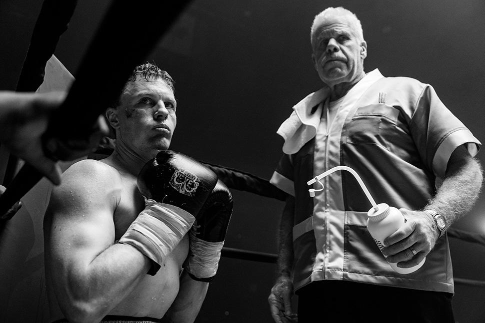 Boxer in ring corner with coach holding water bottle, black and white.