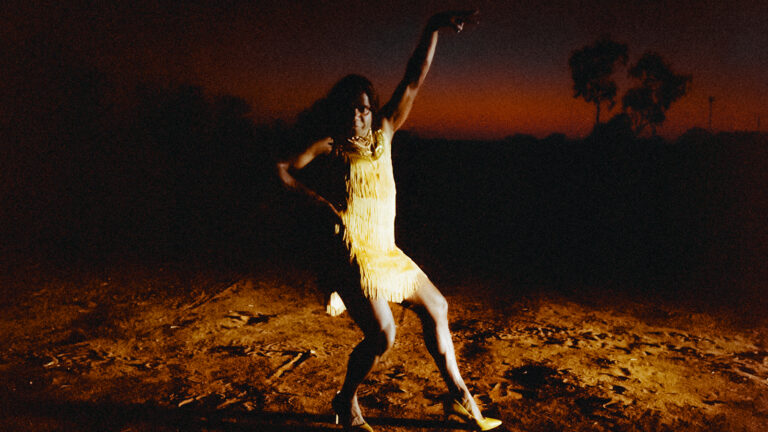 Woman in yellow fringe dress dancing at dusk, Marungka Tjalatjunu (dipped in Black)
