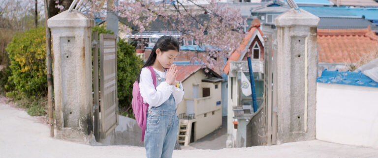 Young girl with backpack praying in a Korean village, cherry blossoms overhead.