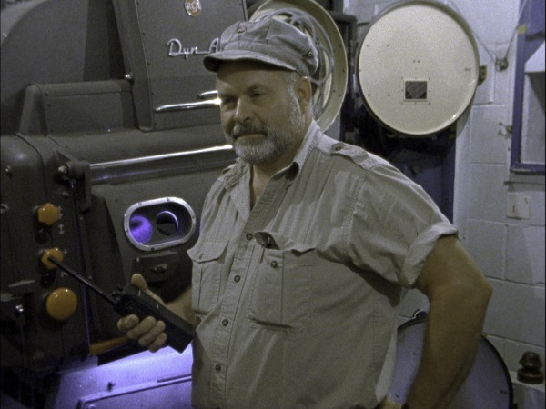 Man in cap with walkie-talkie stands by a Dyn-Arco film projector.
