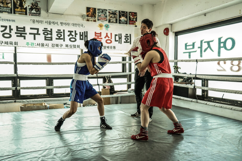 Female boxers sparring in a ring, referee watching closely.