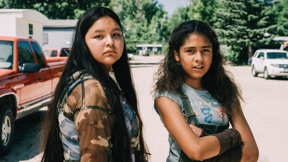 Two Indigenous teenage girls stand outside in a rural setting.