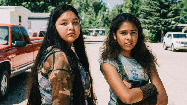 Two Indigenous teenage girls stand outside in a rural setting.