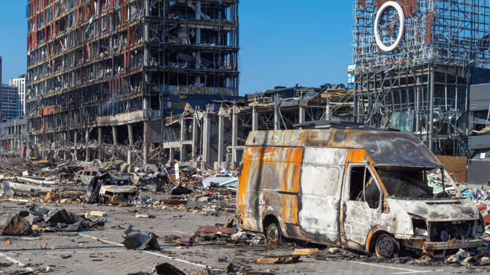 Burnt van amid war destruction in Ukraine. Damaged buildings and debris visible.