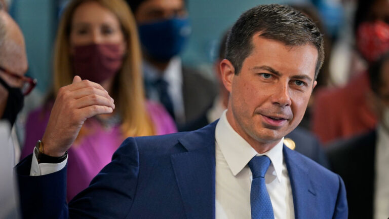 Pete Buttigieg in a blue suit speaks at an event, gesturing with his left hand.