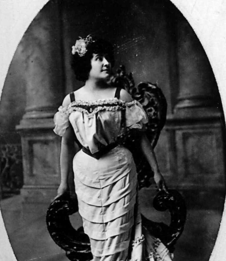 Tekahionwake, Pauline Johnson, in an elegant gown with a flower in her hair, posing by a chair.